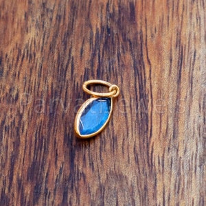 May include: A gold charm with a blue gemstone in a marquise shape. The charm has a loop at the top for attaching to a necklace or bracelet.
