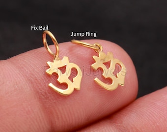 May include: Two small gold-colored Om symbol charms. One charm features a fixed bail at the top, while the other charm is attached to a jump ring. The Om symbol is a recognizable spiritual icon rendered in a polished, metallic gold finish.