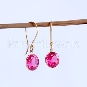 Pode incluir: Um par de brincos de ouro pendurados com pedras redondas cor-de-rosa. Os brincos estão pendurados em um bastão de madeira.