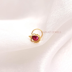 May include: A gold nose ring featuring a teardrop-shaped red stone set in a decorative frame with small gold beads. The jewelry piece is accented with the text PARVATI JEWELS. The design includes a curved wire stem for secure placement.
