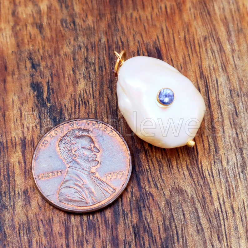 Puede incluir: Un colgante de perla blanca grande, de forma irregular, con una peque&ntilde;a gema azul redonda. El colgante tiene detalles dorados y est&aacute; junto a una moneda para comparar el tama&ntilde;o. El fondo es una superficie de madera oscura.