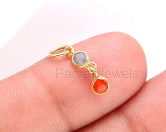Puede incluir: Encanto de oro con un colgante de piedra azul y naranja. El encanto está en un dedo.