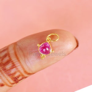 May include: A small gold pendant featuring a pink gemstone set in a triangular shape. The pendant has a loop at the top for attaching to a chain.