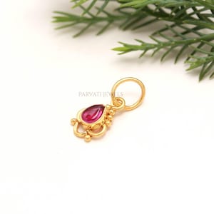 May include: A gold pendant featuring a teardrop-shaped pink gemstone set in a decorative metal frame with small spherical accents and a circular jump ring. The text PARVATI JEWELS is printed in a thin, dark font above the jewellery piece.