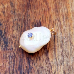 Puede incluir: Un colgante de perla blanca con una fianza dorada y una peque&ntilde;a gema azul redonda. La perla tiene una forma irregular. El colgante est&aacute; sobre un fondo de madera.