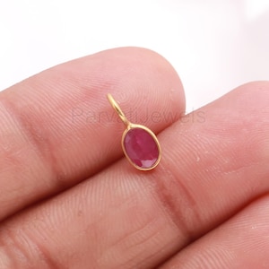 Op de afbeelding: Een kleine, goudkleurige hanger met een ovaalvormige, dieprode edelsteen. De hanger heeft een lusje om aan een ketting of oorbel te bevestigen. De edelsteen is gezet in een gouden zetting.