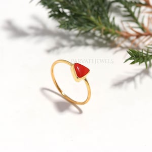 May include: A thin gold ring featuring a small, triangular red stone set in a gold bezel. The ring is positioned upright, and the text PARVATI JEWELS is visible in the background. The design is simple, with a smooth, polished gold band.