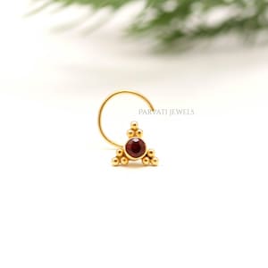 May include: A gold nose ring featuring a triangular cluster design with a central dark red gemstone and small spherical accents. The piece includes the text PARVATI JEWELS. The jewelry has a curved wire post for insertion and a polished finish.