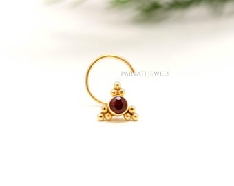 May include: A gold nose ring featuring a triangular cluster design with a central dark red gemstone and small spherical accents. The piece includes the text PARVATI JEWELS. The jewelry has a curved wire post for insertion and a polished finish.