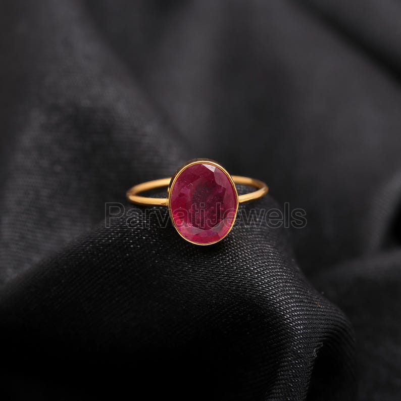May include: A gold ring featuring a large oval ruby gemstone. The ring is displayed against a black fabric backdrop.