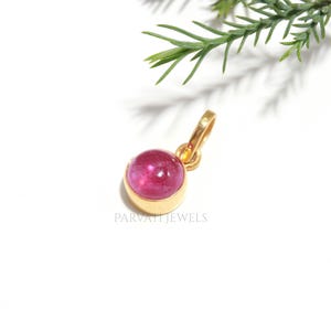 Puede incluir: Un colgante de oro con una piedra preciosa rosa de corte cabujón redondo, engastada en un bisel de metal liso con una pequeña anilla. El texto PARVATI JEWELS está impreso en gris debajo del colgante. El diseño es sencillo y destaca la piedra.