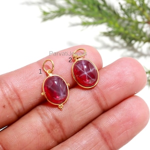 May include: Two gold-colored pendants with oval, deep red gemstones. Each pendant has a star-like pattern within the stone and a small loop at the top for a chain. The pendants are labeled 1 and 2.