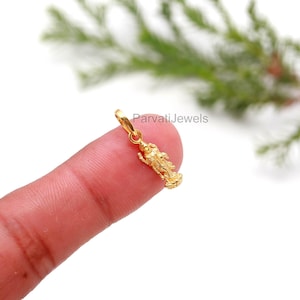 Op de afbeelding: Een kleine, goudkleurige hanger in de vorm van een godheid, vastgehouden tussen vingers. De hanger heeft ingewikkelde details en is opgehangen aan een kleine lus. De tekst "ParvatiJewels" is zichtbaar op de achtergrond.