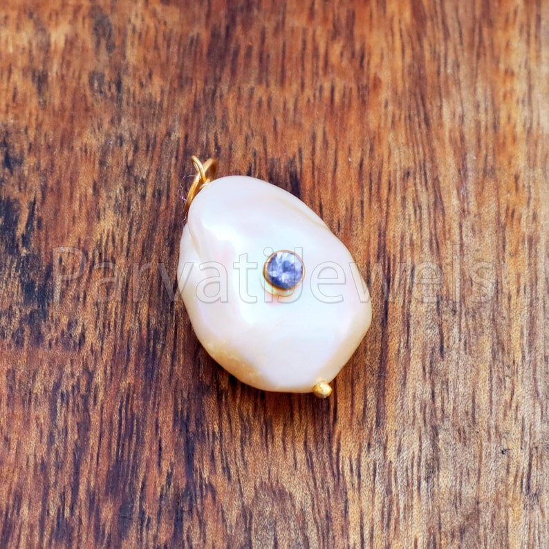 Puede incluir: Un colgante de perla blanca cremosa con una peque&ntilde;a gema azul redonda engastada en el centro. El colgante tiene una argolla dorada y un acento dorado en la parte inferior. El fondo es una superficie de madera.
