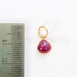 May include: A gold pendant featuring a teardrop-shaped ruby gemstone. The pendant has a small loop at the top for attaching to a chain.