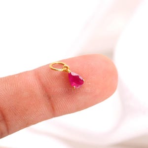 May include: A small teardrop-shaped pendant featuring a vibrant pink gemstone set in a gold-toned metal frame with a circular bail at the top. The text PARVATI JEWELS is visible in faint lettering across the centre of the gemstone surface.