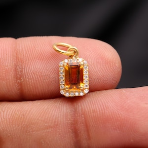 Puede incluir: Un colgante de oro con una piedra preciosa rectangular de color naranja rodeada de pequeñas piedras preciosas transparentes.