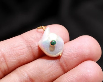 May include: A white, irregularly shaped pearl pendant with a small, round, dark green gemstone set in gold. The pendant has a gold loop at the top for hanging. The background is a dark color.