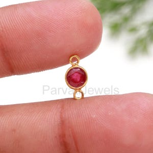 Puede incluir: Conector dorado con una piedra preciosa redonda de color rojo. El conector tiene dos bucles a cada lado.