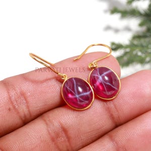 Peut inclure: Une paire de boucles d'oreilles à crochet dorées avec des pierres cabochons ovales rose foncé présentant un effet d'astérisme en forme d'étoile blanche. Le texte PARVATI JEWELS est imprimé en noir près des boucles d'oreilles.