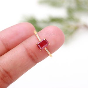 Peut inclure: Bague en or avec une pierre rouge foncé rectangulaire. La fine bande est ornée de petites pierres claires. La bague est tenue par un doigt. Bijou de joaillerie.