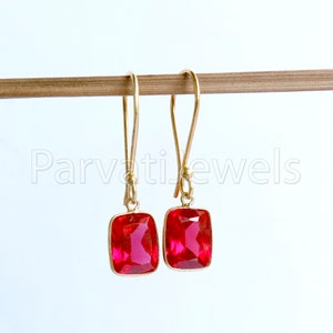 May include: A pair of gold earrings with red square-shaped gemstones. The earrings are hanging from a wooden surface.