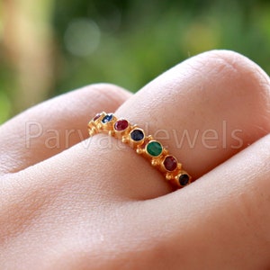 Puede incluir: Un anillo de banda de oro con una fila de piedras preciosas de colores. Las piedras preciosas son rojas, azules, verdes y de un azul más oscuro.