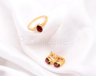 May include: A gold ring and earrings set with oval-shaped red gemstones. The ring and earrings are on a white fabric background.