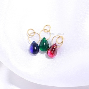May include: Three teardrop-shaped pendants in deep blue, emerald green, and vibrant red, each suspended from a thin gold-toned circular ring. The text PARVATI JEWELS is printed in small black letters near the center of the three jewelry pieces.