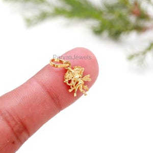 May include: A small, gold-colored pendant in the shape of Cupid, holding a bow and arrow. The pendant is detailed with wings and intricate design. The text "ParvatiJewels" is visible. The pendant is shown on a finger.