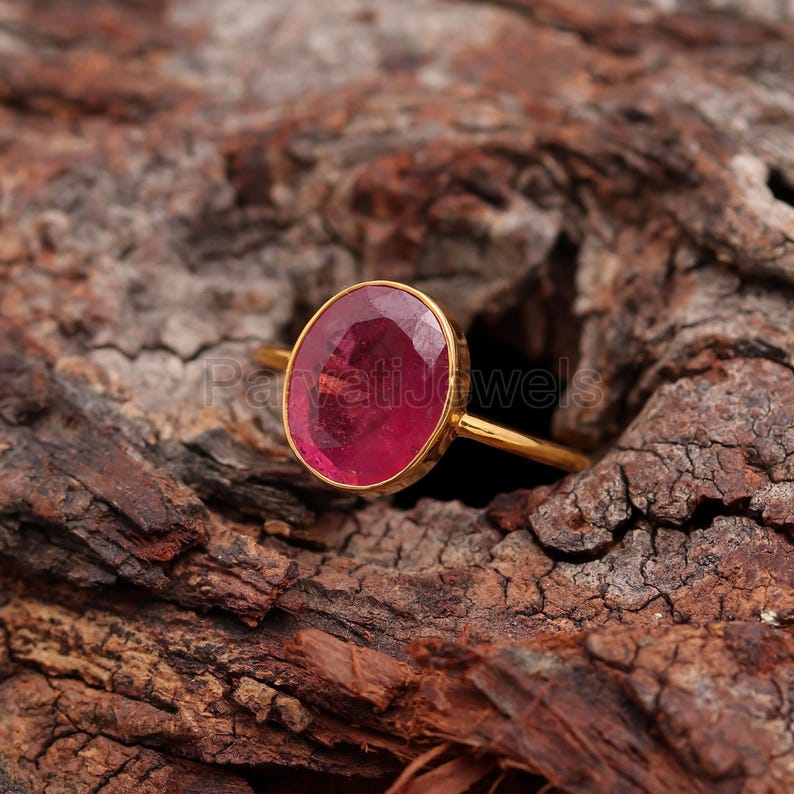 May include: A gold ring featuring a large, oval-shaped, pink gemstone. The ring is displayed on a wooden surface.