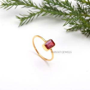 Puede incluir: Un anillo de oro con una piedra preciosa roja rectangular facetada engastada en una banda de oro sencilla. El anillo lleva el texto PARVATI JEWELS. El diseño consiste en una banda fina de hilo de oro pulido que sostiene una piedra roja.