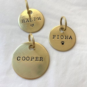 May include: Three gold-colored metal dog tags with engraved names. The tags are round and have a small ring attached to the top. The names on the tags are "Ralph" with a heart symbol, "Fiona" with a paw print symbol, and "Cooper".