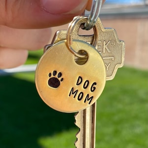 May include: A gold-colored key chain with a round tag that says "DOG MOM" and has a paw print. The tag is attached to a key with a gold-colored key ring.