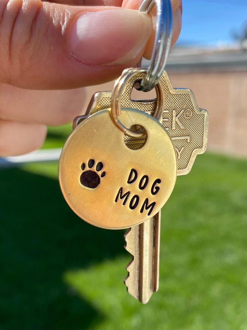 May include: A gold-colored key chain with a round tag that says "DOG MOM" and has a paw print. The tag is attached to a key with a gold-colored key ring.
