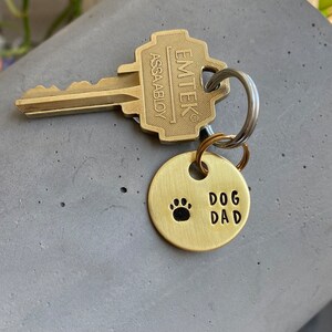 May include: A gold-toned key chain with a round tag that says "DOG DAD" with a paw print. The key chain is attached to a silver key with a brass key head that says "EMTEK ASSA ABLOY".