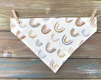 May include: A white bandana with a pattern of colorful rainbows. The bandana is tied with a piece of twine and two wooden clothespins.