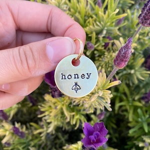 May include: A gold-coloured metal tag with the word "honey" engraved on it. A small bee design is also engraved on the tag.