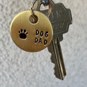 May include: A gold-colored metal keychain with a round tag that says "DOG DAD" and has a paw print. The keychain is attached to a silver key.