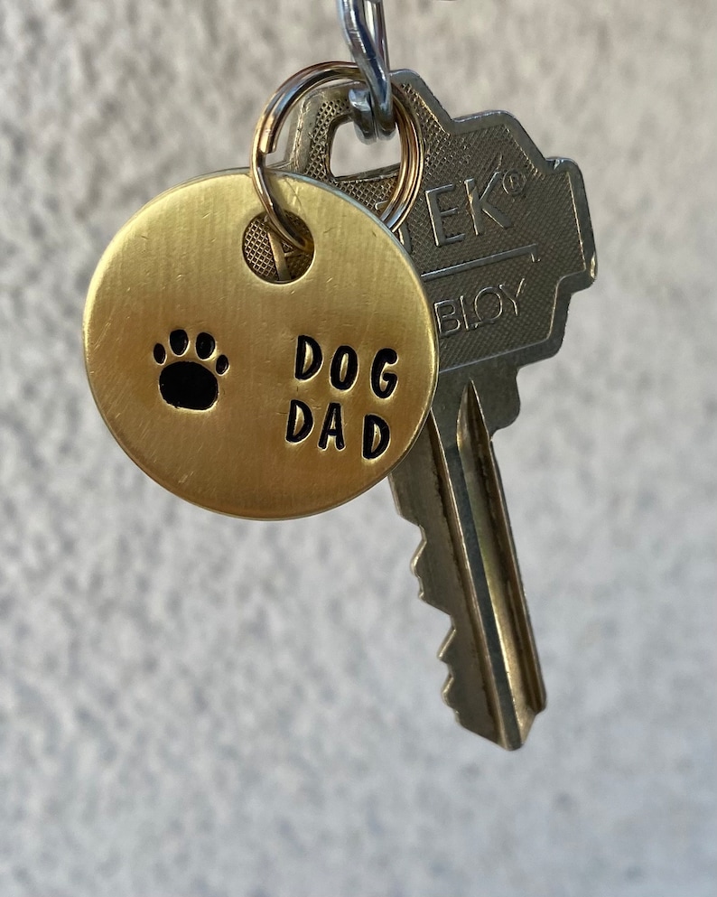 May include: A gold-colored metal keychain with a round tag that says "DOG DAD" and has a paw print. The keychain is attached to a silver key.