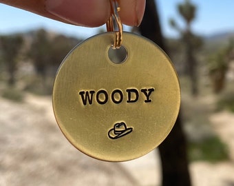 May include: A gold-colored metal tag with the word "WOODY" engraved in black lettering. A small, engraved image of a cowboy hat is below the text.