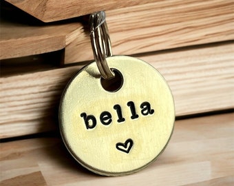 May include: A round, brass-colored pet tag with the name "bella" in black, lowercase letters and a small heart symbol. The tag is attached to a silver-colored split ring. The background is a wooden surface.
