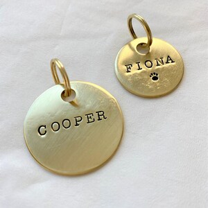 May include: Two gold-colored metal dog tags with engraved names. The tag on the left says "COOPER" and the tag on the right says "FIONA" with a paw print symbol.