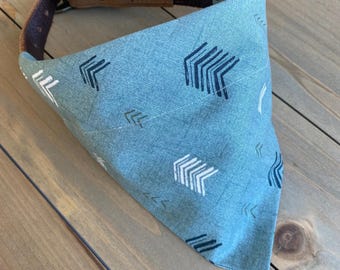 May include: A light blue dog bandana with a brown leather collar. The bandana has a pattern of white and black arrows.