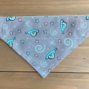 May include: A gray dog bandana with a blue and green alien spaceship pattern. The bandana is decorated with pink and white stars and swirls.
