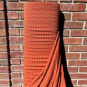 May include: A roll of orange textured fabric with a honeycomb pattern. The fabric is folded and draped over a brick wall.