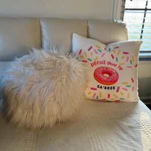 May include: A white, fluffy, round pillow and two decorative pillows on a white surface. One pillow has a pink donut graphic with the text "donut grow up" and "SA'BREE". The other pillow is white.