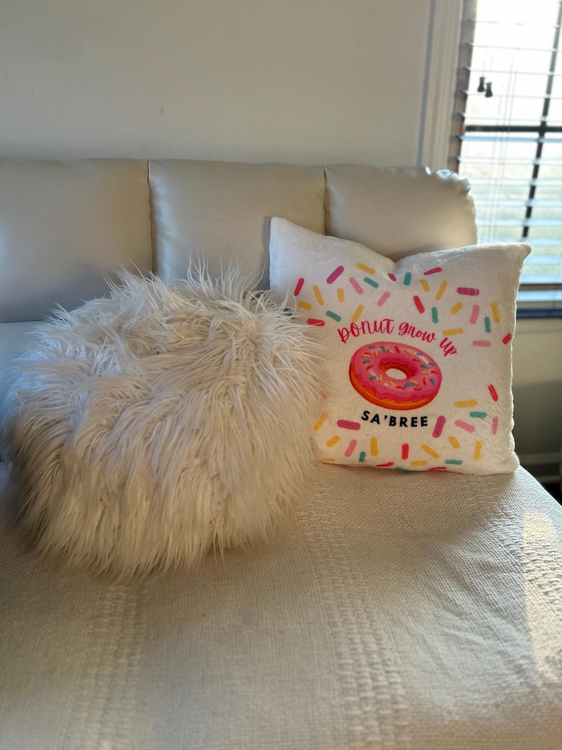 May include: A white, fluffy, round pillow and two decorative pillows on a white surface. One pillow has a pink donut graphic with the text "donut grow up" and "SA'BREE". The other pillow is white.