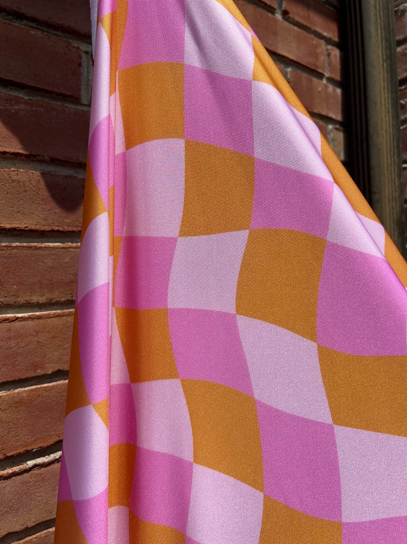May include: A pink and orange checkered fabric with a wavy pattern. The fabric is folded and draped over a brick wall.