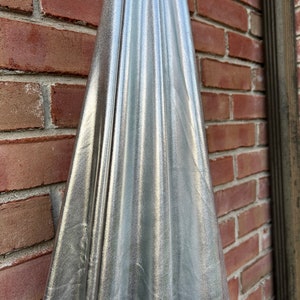 Könnte beinhalten: Ein silberner Metallic-Stoff mit glatter, glänzender Oberfläche. Der Stoff ist über eine Backsteinmauer drapiert und bildet Falten und Knitter.
